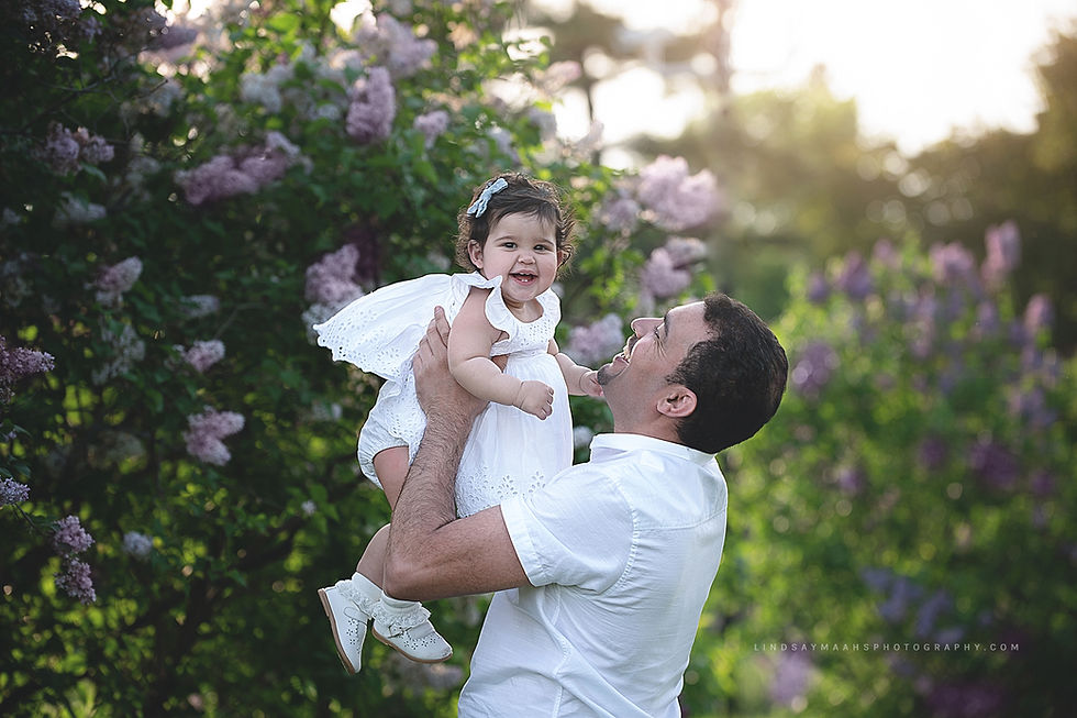 Portrait of dad holding baby girl up in the air. Baby and dad are both laughing among the lilacs