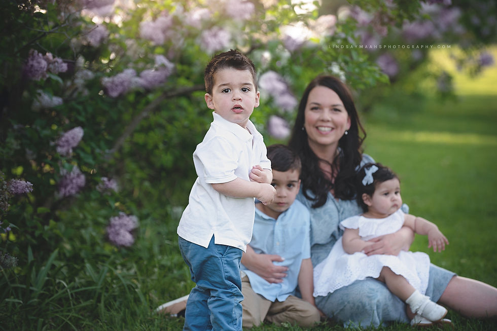 Portrait of a Mom sitting on grass in front of lilacs holding her daughter. One son is sitting next to her and the other standing closer to the camera.