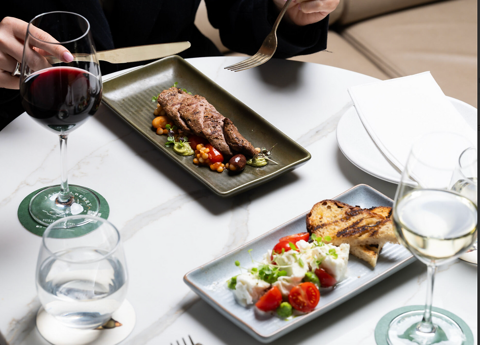 Hands holding utensils over a dish with meat and vegetables on a marble table, paired with red and white wine, evoking a sophisticated dining scene.