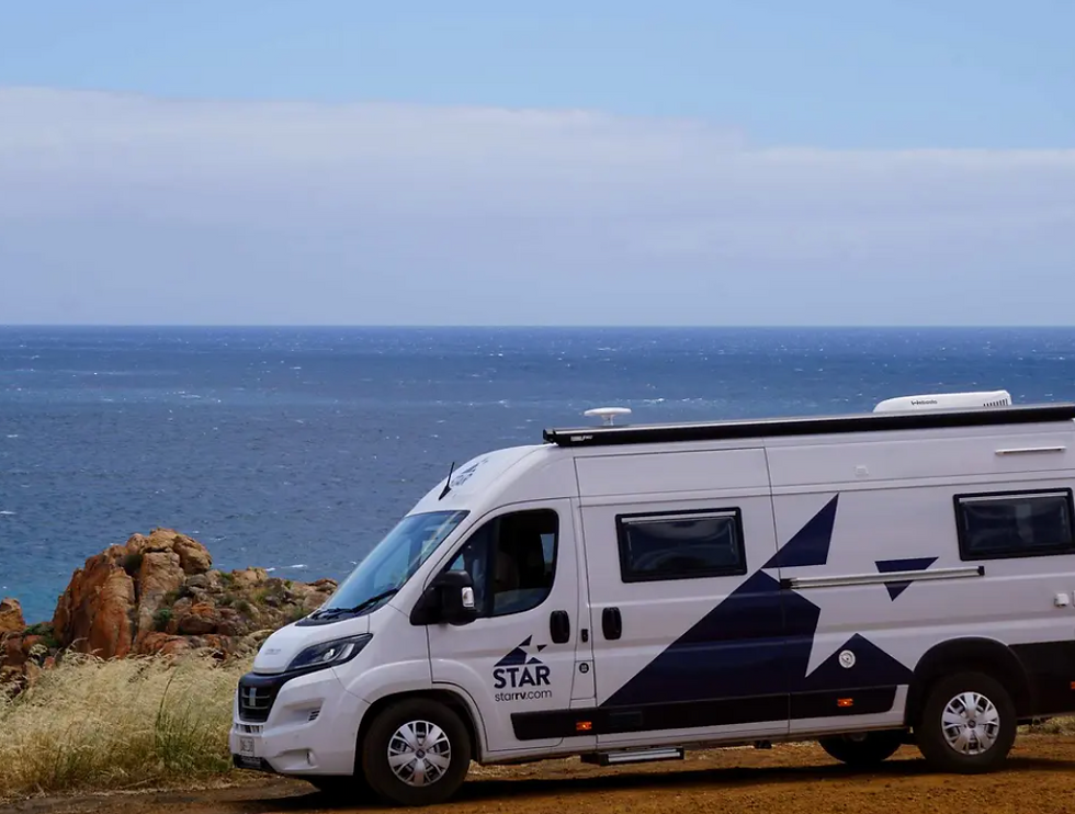 White STAR camper van parked on a coastal dirt road beside rocky cliffs and a calm blue ocean under a clear sky.