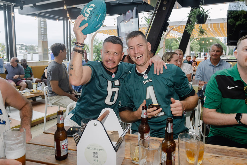 Two men in green jerseys cheer in a bar, one holding a football. Smiling, surrounded by drinks and people. Bright and lively atmosphere.