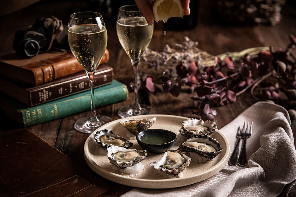 Plate of oysters with lemon juice being squeezed over them, two glasses of champagne, books, and dried flowers on a wooden table at the Botanist Sydney