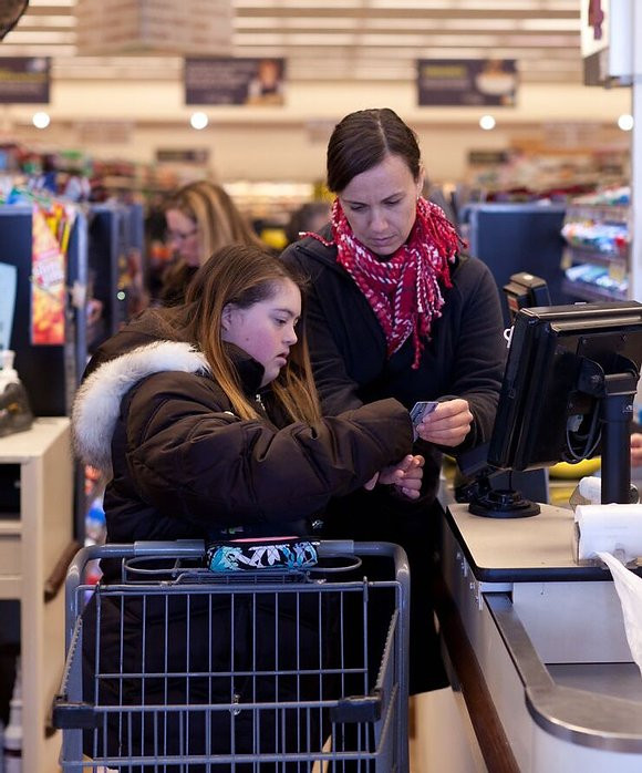 Image of Danielle Kelly, wearing a pink scarf and black jacket, assisting student Emma as she starts to pay for her purchase at the grocery store.