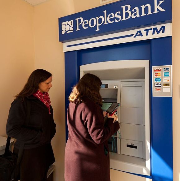 Danielle Kelly, wearing a pink scarf and black jacket, looks on as student Carly uses the PeoplesBank ATM.