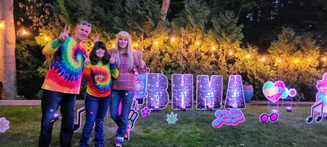 Image of Joe, Paige, and Karen Phillips dressed in colorful tie dye posing outside in front of BHMA lawn decorations.