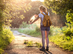 Woman sprays mosquito repellent on arm in sunny forest path, wearing a yellow backpack. Golden sunlight filters through green foliage.