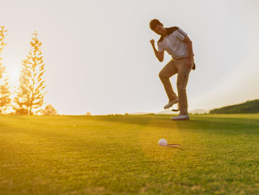 Golfer fist pumps in excitement on sunny course. Golf ball on green grass, trees in the background, warm sunset glow enhances joyful mood.