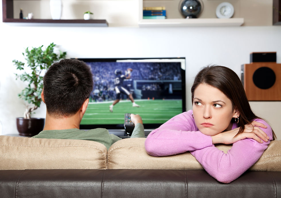 Man watching football on TV, sitting on a couch beside a woman in a pink sweater looking bored. Living room setting with shelves.