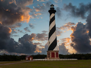 Cape Hatteras Lighthouse – North Carolina. Lighthouse with black and white stripes stands against a dramatic sunset sky, with dark clouds and a small red-brick building nearby.