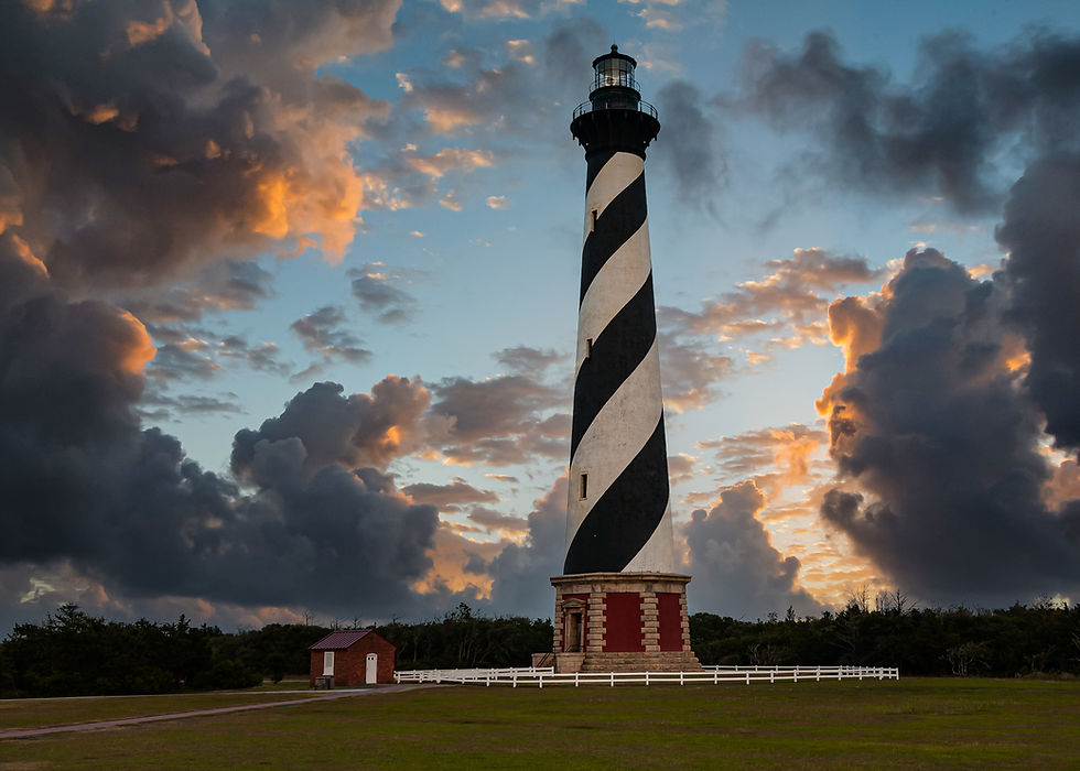 Cape Hatteras Lighthouse – North Carolina. Lighthouse with black and white stripes stands against a dramatic sunset sky, with dark clouds and a small red-brick building nearby.