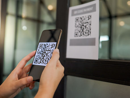 Hands holding a smartphone scanning a QR code on glass. Background shows a paper sign with QR code and "QR CODE PAYMENT" text. Bright setting.
