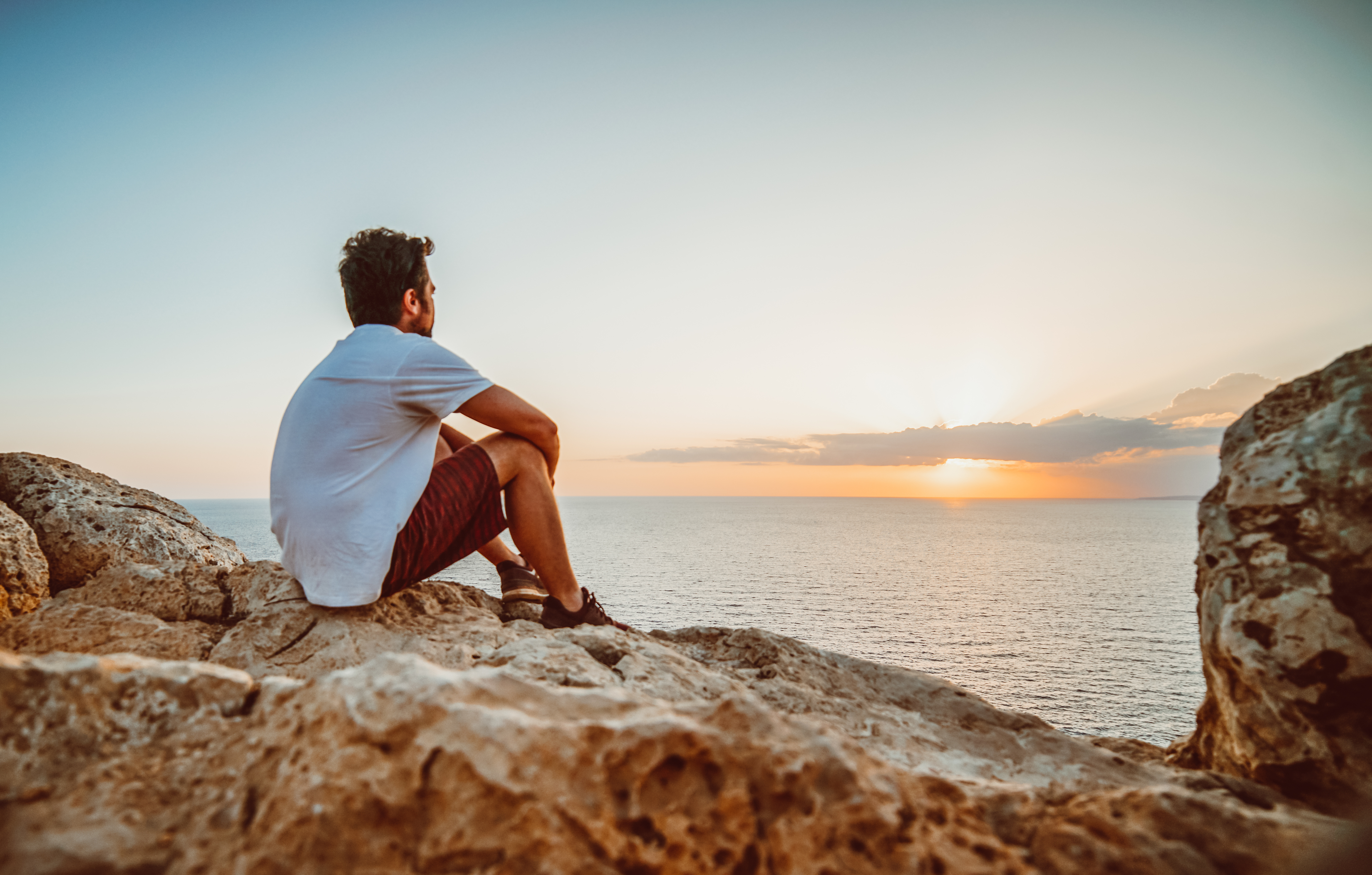 Man in white T-shirt sits on rocky cliff, gazing at sunset over the ocean. Sky is clear with warm orange hues, creating a calm mood.