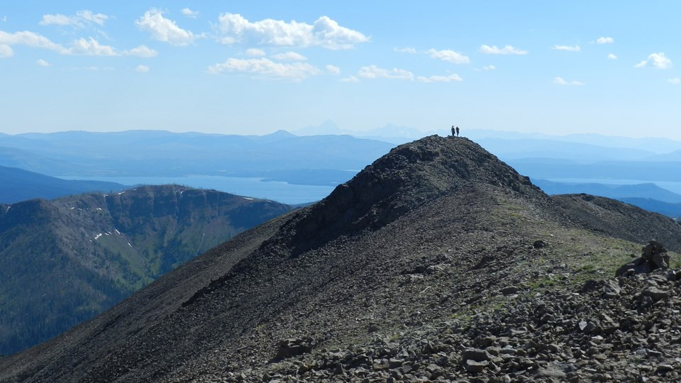 Two people atop the summit of rocky Avalanche Peak in Yellowstone National Park, looking out over an incredible view