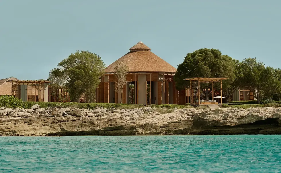 Circular wood building with conical roof by turquoise sea. Surrounded by trees and rocky shore, clear sky background, peaceful ambiance.