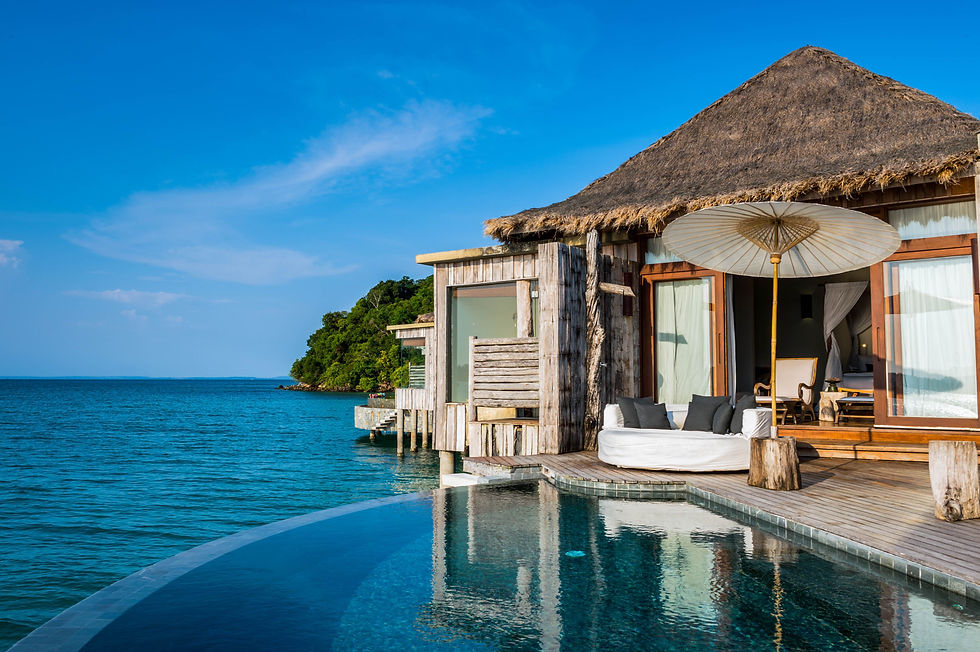 Villa with thatched roof by a blue ocean, featuring a pool, white sofa, and parasol on wooden deck. Tropical setting, clear sky, lush greenery.