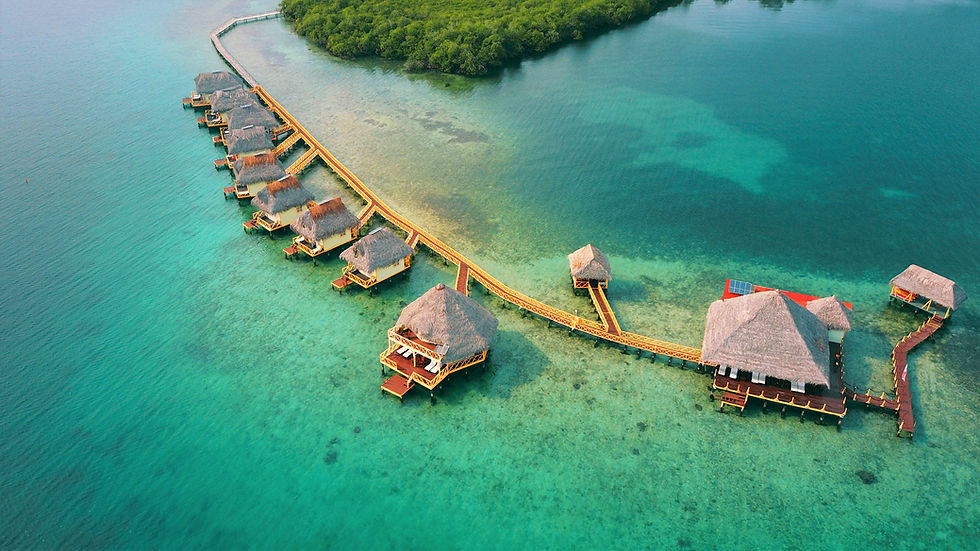 Overwater bungalows with thatched roofs connected by wooden walkways, set over clear turquoise water, surrounded by greenery.