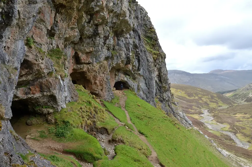 Steep rocky cliff with caves, a narrow grassy path leads inside. Mountain landscape in the background under a cloudy sky.