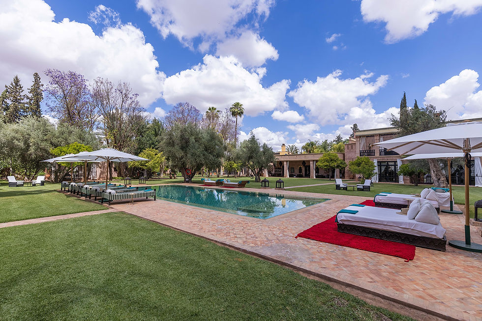 Luxurious poolside setting with loungers, umbrellas, and a lush garden. Clear blue sky and fluffy clouds overhead create a serene, inviting atmosphere.