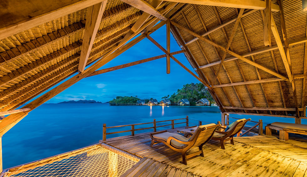 Ocean-view deck at sunset with wooden lounge chairs under a thatched roof, overlooking serene blue waters and distant island huts.