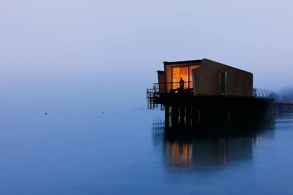 A person stands on a lit balcony of a cabin over calm water, with a misty blue background creating a serene atmosphere.