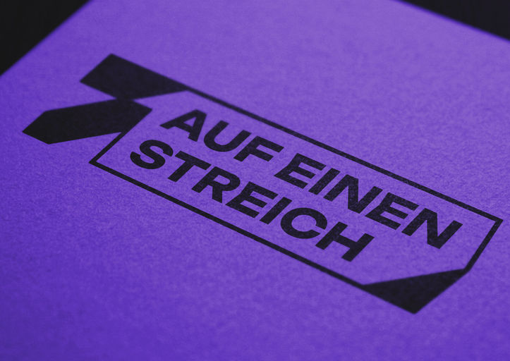 Close-up of 7 auf einen Streich logo printed on purple surface.