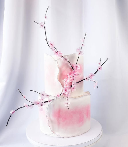 Minimalist two-tier white wedding cake with soft pink watercolor accents and sculptural branches covered in delicate pink cherry blossoms.