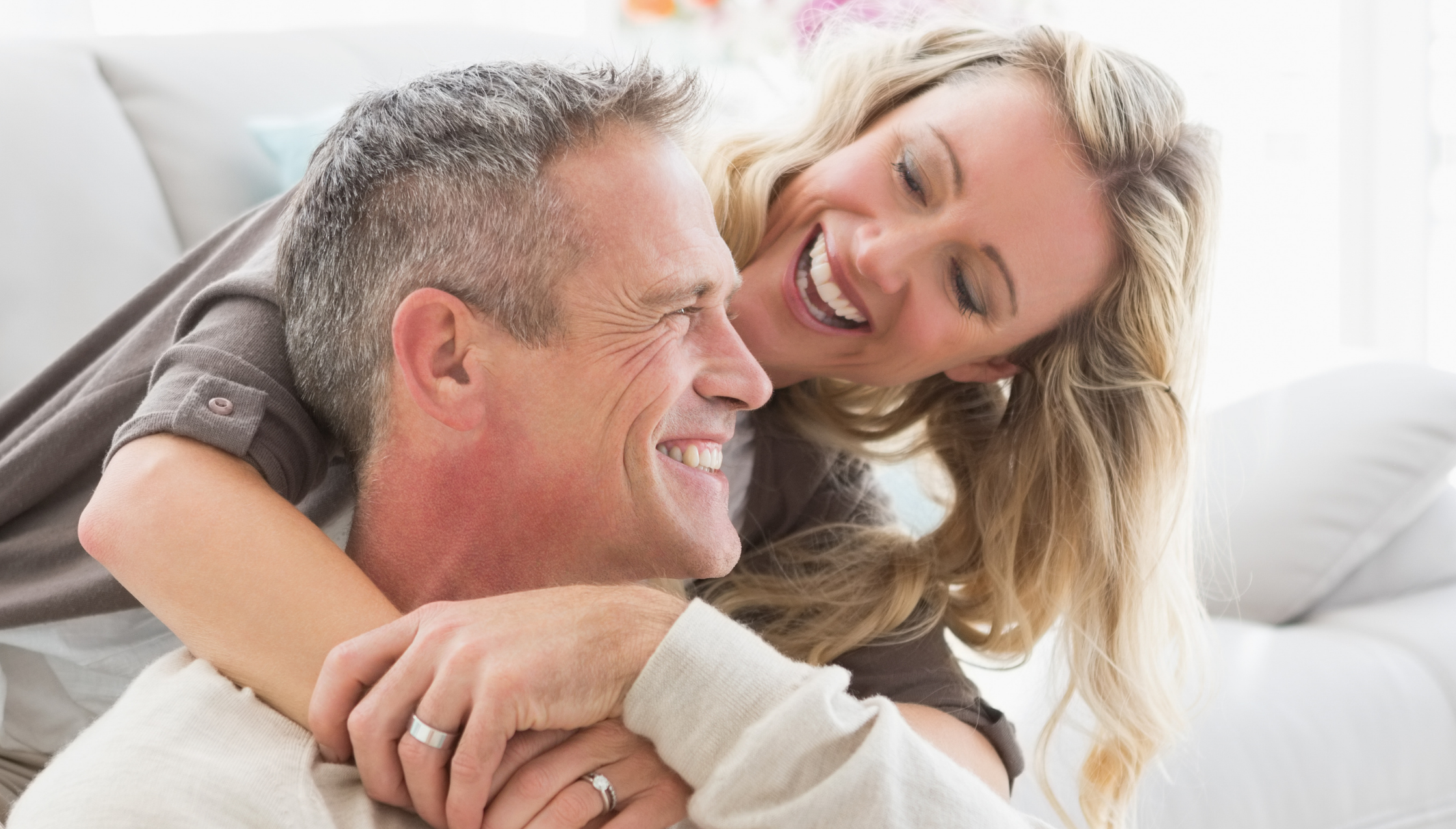Smiling couple hugging on a bright white couch in a sunlit living room, sharing a warm, affectionate moment.