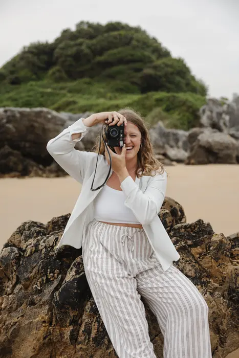 Am Strand lehnt sich die Fotografin in Stuttgart an einem Stein an und schaut durch ihre analoge Kamera hindurch und lächelt.