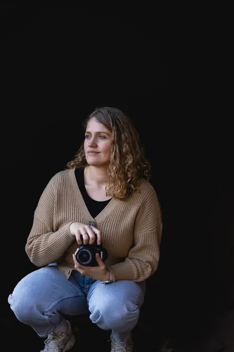 Junge Fotografin kniet in der Hocke vor einem schwarzen Hintergrund. Wie es sich für ein Fotograf gehört, hält sie die Kamera in der Hand. 
