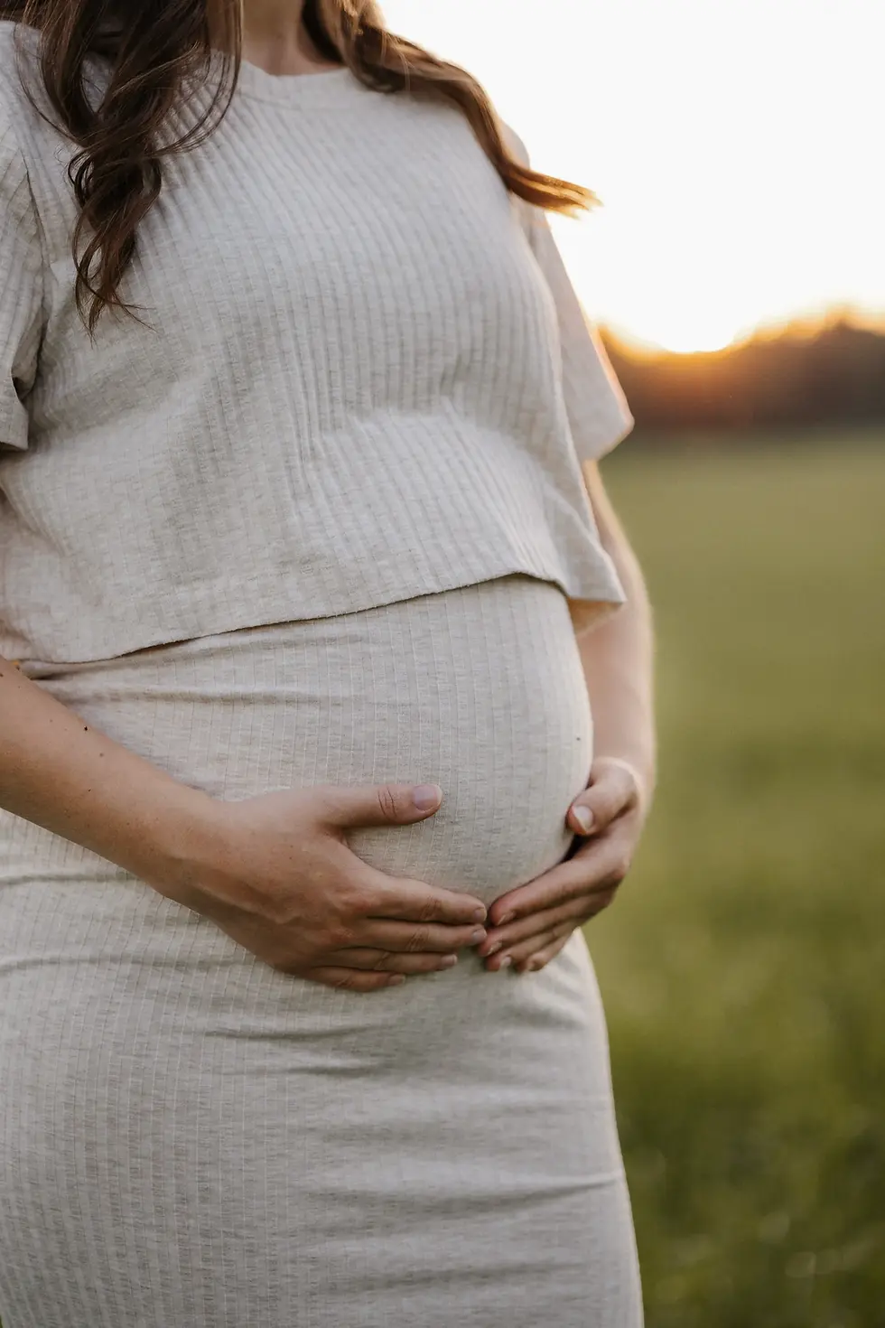 Babybauchshooting bei Sonnenuntergang. Sie hält mit beiden Händen den Bauch, man sieht kein Gesicht.
