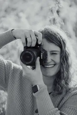 Schwarz weiß Foto von einem Fotograf. Sie ist weiblich und lächelt während sie durch ihre eigenen Kamera blickt. 
