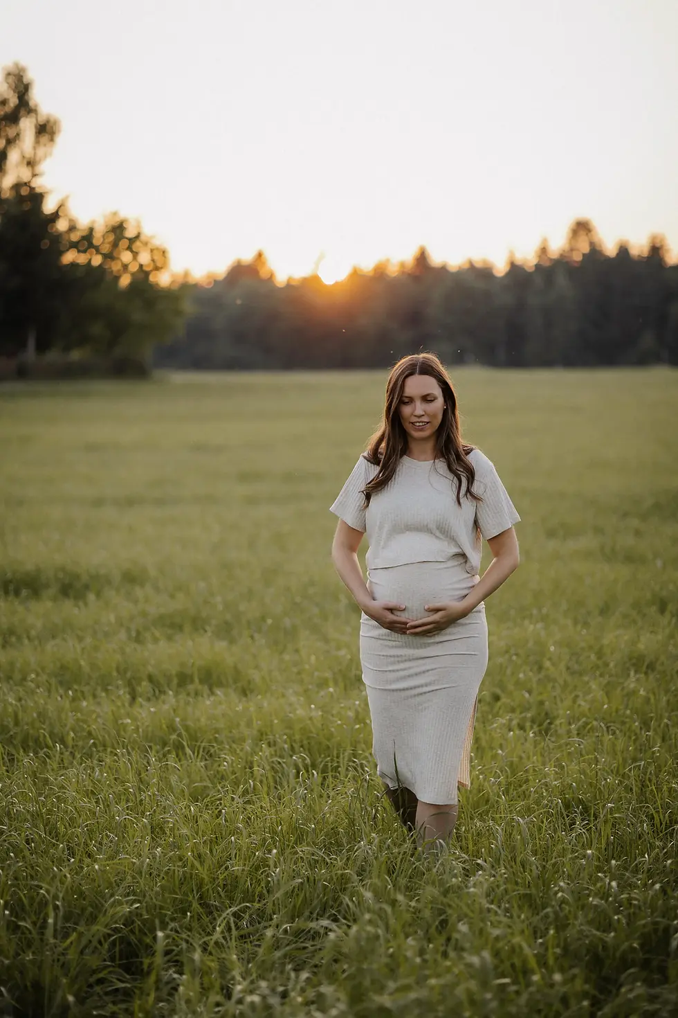 Babybauchshooting auf einer Wiese bei Sonnenuntergang. Sie hält beide Hände an den Bauch und schaut herunter.