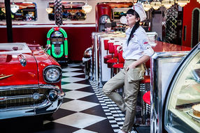 An American diner scene featuring a classic Chevy car, complete with staff members blowing giant chewing gum balloons, evoking a nostalgic 1950s ambiance.