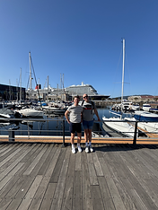 Travel content creators Tom and Dom of Tom and Dom Travel standing at the Port of Vigo in Galicia, Spain, with the luxury Cunard cruise liner Queen Anne docked prominently in the background showing its iconic black, white, and red hull.