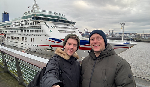 Tom and Dom from Tom and Dom Travel standing together outside the modern glass building of the Altona Cruise Terminal in Hamburg, with the white P&O Aurora cruise ship docked in the background on the River Elbe.