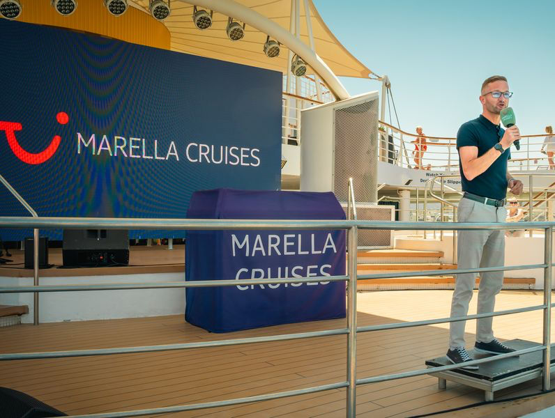 Scott Sanders Cruise Director speaking into a microphone on a cruise ship stage beside Marella Cruises signage, with railings and sunlit deck behind.