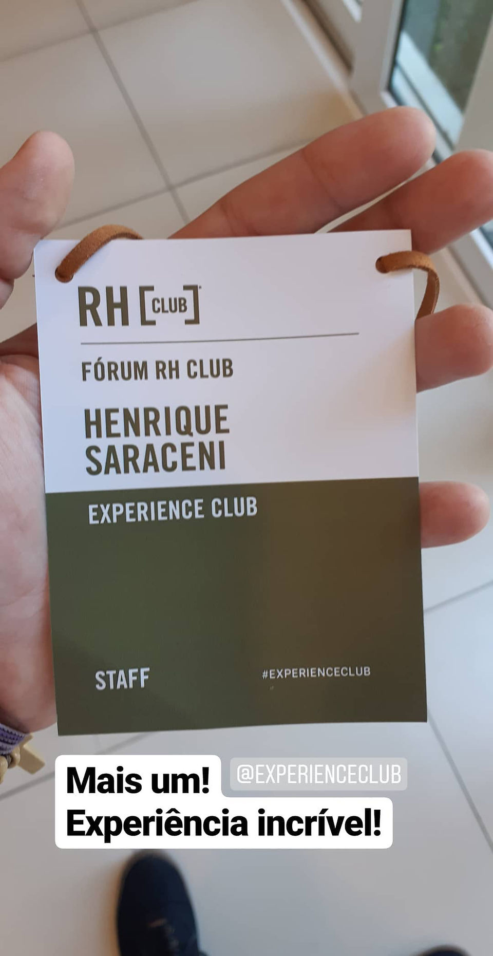 Foto vertical capturada da perspectiva em primeira pessoa, mostrando uma mão aberta segurando um crachá preso a um cordão marrom claro sobre um piso claro. A parte superior do crachá é branca com 'RH [CLUB]', 'FÓRUM RH CLUB' e o nome 'HENRIQUE SARACENI'. A parte inferior é verde-oliva com 'EXPERIENCE CLUB', 'STAFF' e '#EXPERIENCECLUB'. Na base da imagem, há duas caixas de texto sobrepostas: 'Mais um! @EXPERIENCECLUB' e 'Experiência incrível!'.