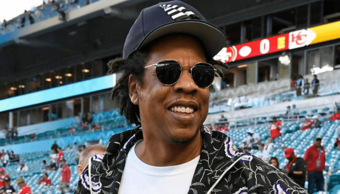 Das Bild zeigt Jay Z in einem Basballstadion. Er trägt ein schwarzes Hawaii-Hemd, Basecap und eine Sonnenbrille
