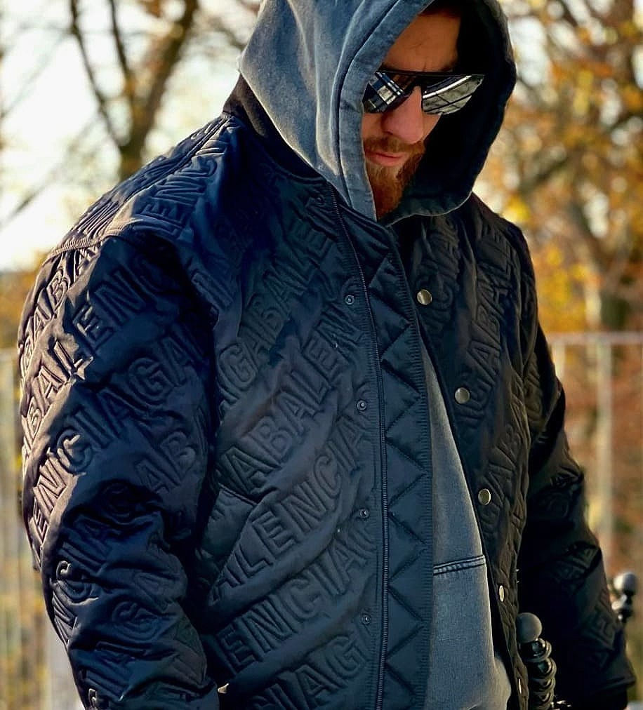 Das Bild zeigt Entertainer Mois in einem Park. Gelbe Blätter sind an den Bäumen zu sehen. Mois trägt einen grauen Hoodie mit Kapuze über dem Kopf. Darüber eine schwarze Jacke und schwarze Sonnenbrille mit silber Gläsern.