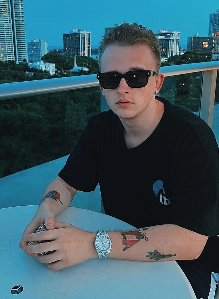 Das Bild zeigt Producer Pvlace. Er sitz auf einer Terasse in einem höherem Stockwerk. Im Hintergrund seit mehrere Wolkenkratzer zu sehen. Pvlace trägt ein schwarzes T-Shirt und eine schwarze Sonnenbrille. Am Arm trägt er eine silberne Uhr.