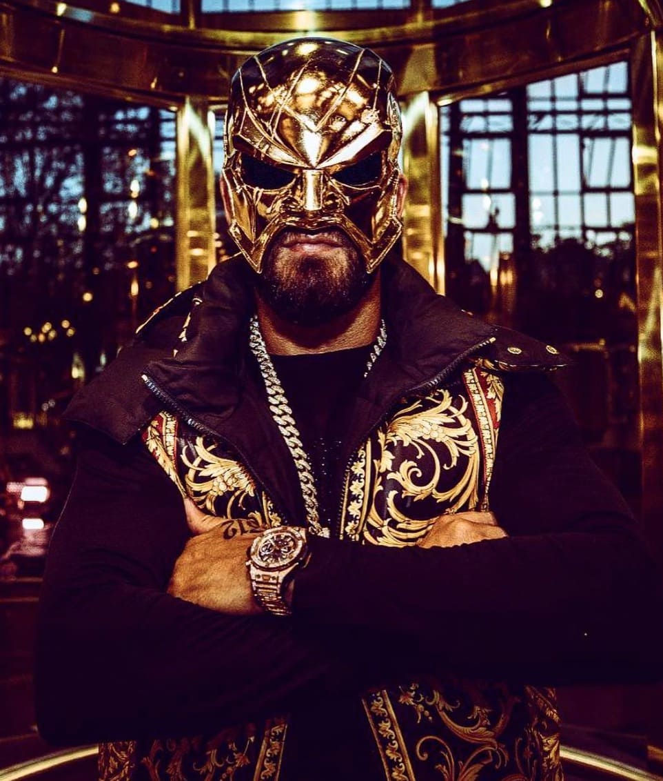 Das Bild zeigt Rapper 18 Karat mit einer goldenen Maske vor seinem Gesicht. Er trägt eine schwarz goldene Jacke, eine Silberkette und eine silberne Uhr am Handgelenk.
