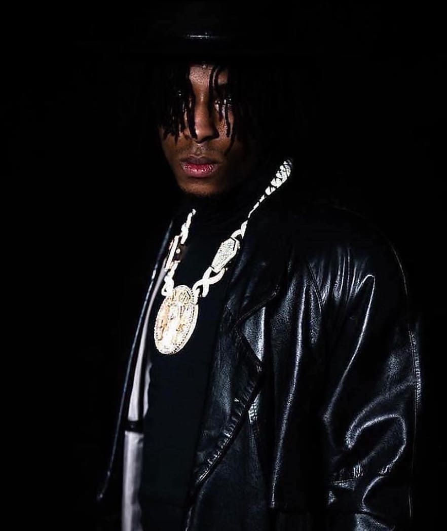 Das Bild zeigt Rapper NBA Youngboy vor schwarzem Hintergrund. Der Rapper trägt einen schwarzen Ledermantel und eine schwarze Mütze. Um den Hals trägt er eine Silberkette.