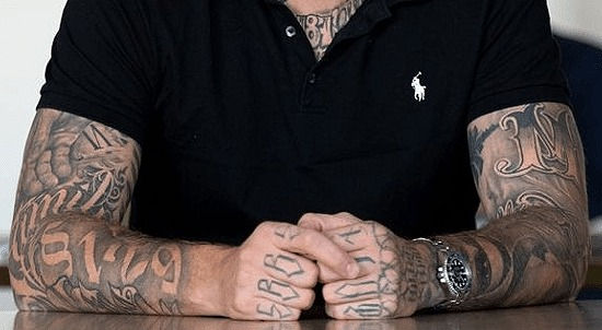 Das Bild zeigt Oberkörper und Arme des Rappers Gzuz. Ein schwarzes T-Shirt der Marke Ralph Laurent ist zu sehen, sowie beide Arme und Hände komplett tätowiert.