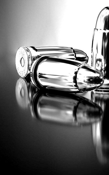 Das Bild zeigt drei Patronen in schwarz weiß, die von einer Pistole stammen und auf einem Tisch liegen. Das Foto ist Metallic glänzend und spiegelt die Patronen auf dem Tisch wieder.