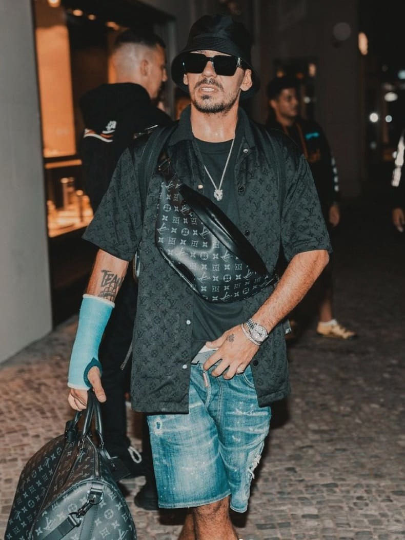 Das Bild zeigt den Rapper Capital Bra auf der Straße. Im Hintergrund sind ein paar Menschen zu sehen. Capital Bra trägt ein schwarzes Luis Vuitton Hemd, Bauchtasche am Oberkörper und in der rechten Hand ein schwarze Luis Vuitton Reisetasche. Dazu eine blaue Jeans Short und eine schwarze Sonnenbrille und Basecap. Am rechten Arm hat er einen blauen Gipsverband