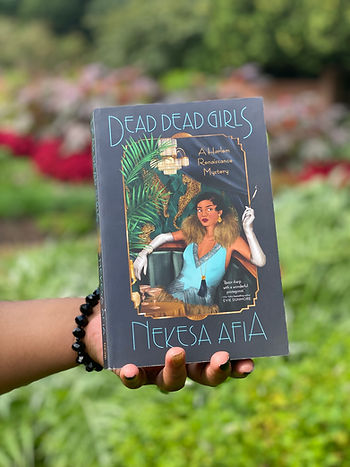 My hand holding a copy of Dead Dead Girls in front of a green and red garden.