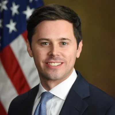 Paul Perkins smiles in a dark suit while sitting in front of an American flag.