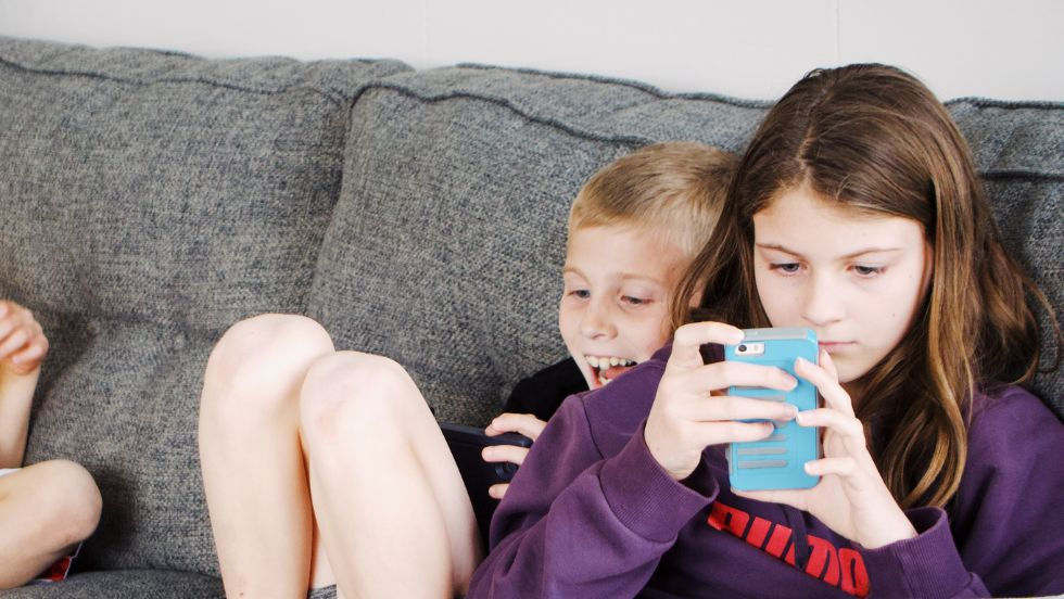 Two children sat on a grey couch, looking at smart phones. One child is smiling and one is looking serious at their blue smartphone.