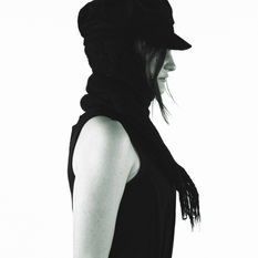 A white high-fashion model stands at a profile angle. The background is entirely white and we only see her silhouette wearing a tight-fitting black dress, a black scarf, and a black hat.