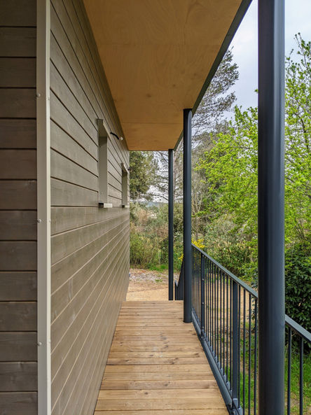 Balcon en bois long et étroit longeant la façade d'une maison avec un bardage en bois. La couverture du balcon est soutenue par des poteaux métalliques noirs, offrant une structure minimaliste et élégante. Le design de l'espace met en valeur l'intégration avec la nature environnante, avec une vue sur la végétation à travers les garde-corps en métal. Le mariage des matériaux, entre bois naturel et métal, crée une atmosphère moderne tout en restant en harmonie avec l'environnement extérieur.